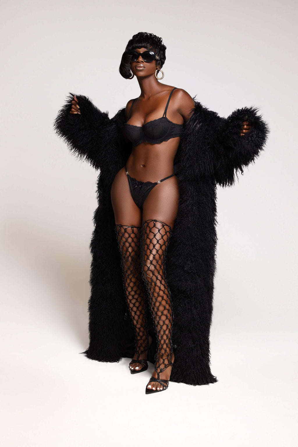 Model showcasing a luxurious black Mongolian fur trench coat with sultry lingerie and thigh-high fishnet stockings.